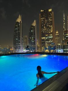 a woman sitting in the middle of a swimming pool at night with skyscrapers in the background