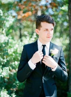 a young man in a suit and tie standing next to trees with his hands on his lapel