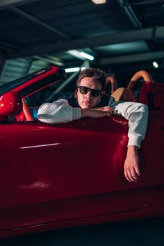 a man in sunglasses leaning on the hood of a red car with his arms crossed