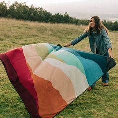 a woman holding a large multicolored blanket on top of a lush green field