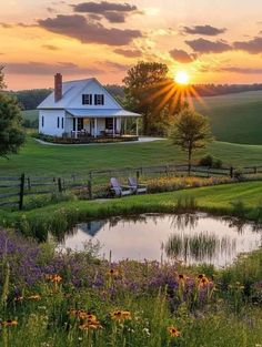 the sun is setting over a farm house with a pond and flowers in front of it