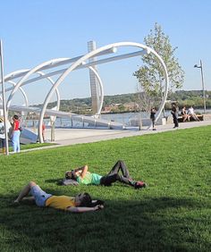 two people laying on the grass in front of a bridge and some buildings with water behind them