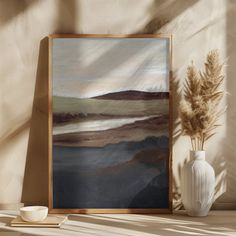 an abstract painting in a wooden frame on a shelf next to a vase with dried grass