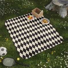 a black and white checkered picnic blanket on the grass with tennis rackets, fruit and bread