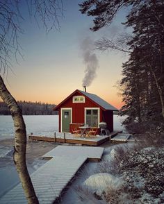 a small red cabin sitting on top of a snow covered ground next to trees and water