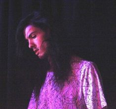 a man with long black hair standing in front of a microphone and wearing a purple shirt