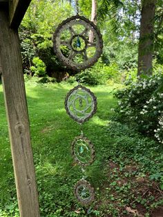 three metal wind chimes hanging from a wooden post in the grass with trees and bushes behind them