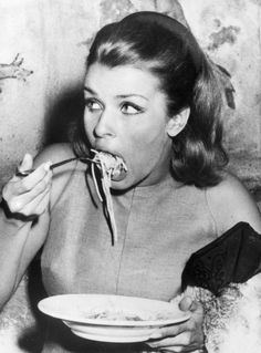 a black and white photo of a woman eating noodles with chopsticks in her mouth