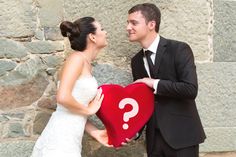 a bride and groom holding a heart shaped pillow with a question mark on it while standing next to a stone wall