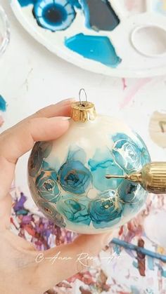 a hand holding a blue and white ornament in front of paintbrushes