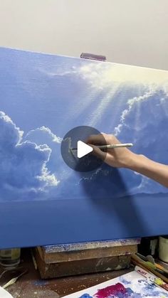 a person is painting clouds in the sky on a blue canvas with an easel