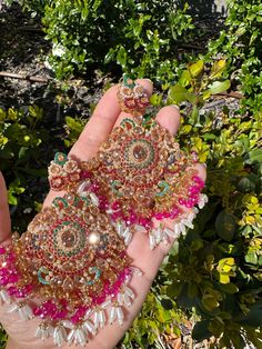 a hand holding two pairs of earrings in front of some bushes and shrubbery with green leaves