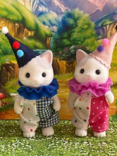 two white kittens wearing colorful outfits and hats are standing next to each other in front of a painting