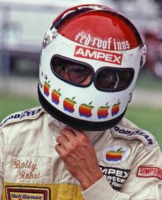 a man in a racing suit and helmet holding his hand up to his face while standing on a field