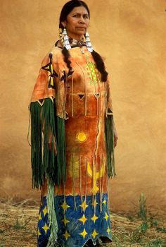 a native american woman standing in front of a tan wall wearing an orange and blue dress