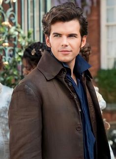 a man standing in front of a building wearing a brown coat and blue dress shirt