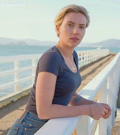 a woman leaning on a white fence by the ocean with her hand on her hip