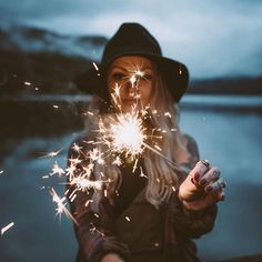 a woman is holding sparklers in her hands and looking at the camera while standing near water