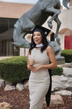 a woman in a dress and hat standing next to a horse statue holding a drink