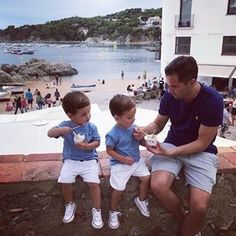 a man sitting next to two young boys eating ice cream at the beach while others watch