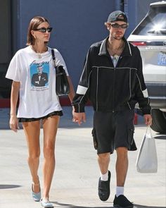 the man and woman are walking down the street holding hands while wearing black shorts, white t - shirt and sneakers