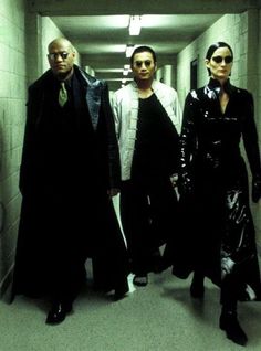 three people walking down a long hallway in black clothes and white shirts, with one person wearing