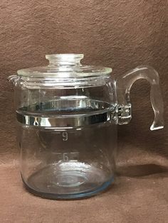 a clear glass tea pot with a lid on a brown cloth covered tableclothed surface