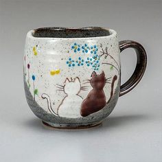 a coffee cup with two cats on it and flowers painted on the side, sitting in front of a gray background