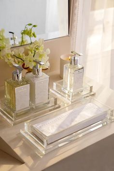 two clear glass containers with perfumes on a table next to a mirror and flowers