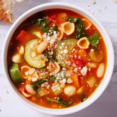 a white bowl filled with pasta and veggies soup next to a slice of bread