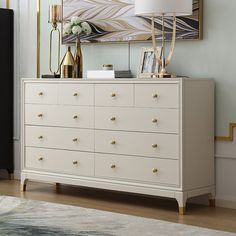 a white dresser with gold handles in front of a painting on the wall and lamp