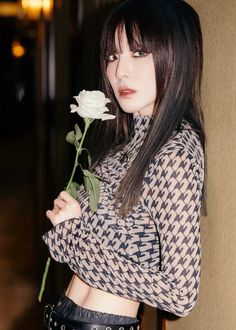 a woman holding a white rose in her right hand and looking at the camera while standing next to a wall