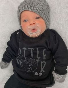 a baby with a pacifier in his mouth laying on a white blanket wearing a sweater and hat