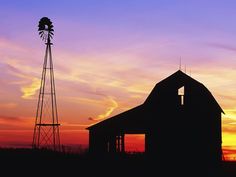 a barn and windmill at sunset with the sun setting in the sky behind it,