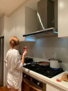 a woman standing in a kitchen holding a glass of wine and looking at the stove