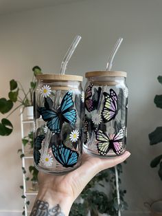 two butterflies painted on glass mason jars with straws in them, held by a person's hand