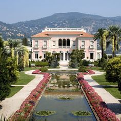 a large pink house surrounded by lush green trees and flowers with a fountain in the foreground