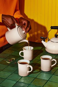 a person pours coffee into three mugs on a green tile table with yellow walls