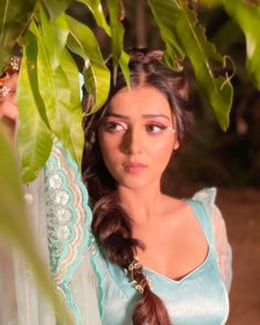 a woman with long hair wearing a blue dress standing under a leafy tree and looking at the camera