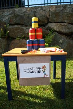 a wooden table topped with lots of different colored cups on top of green grass next to a rock wall