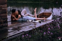 a man and woman laying on a wooden dock next to a lake with a boat in the water