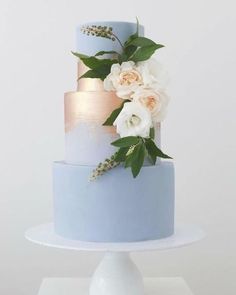 a three tiered blue cake with white flowers and greenery on the top layer