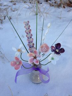 an arrangement of flowers in a glass vase on the snow covered ground with purple ribbon