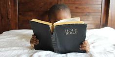 a young boy reading a bible on top of a bed