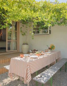 the table is set outside with plates and cups on it, near a bench under a tree