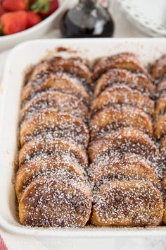 powdered sugar covered pastries in a white baking dish with strawberries on the side