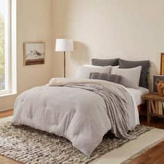 a bed with white sheets and grey pillows in a bedroom next to a large window