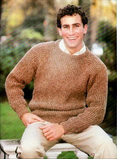 a man sitting on top of a wooden bench wearing a brown sweater and khaki pants