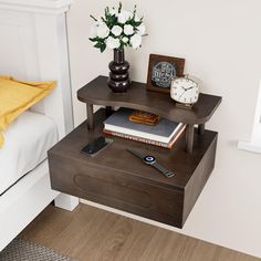 a nightstand with a phone, books and flowers on it next to a white bed