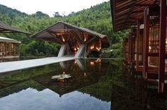 an outdoor swimming pool surrounded by wooden buildings and greenery at dusk, with the lights on